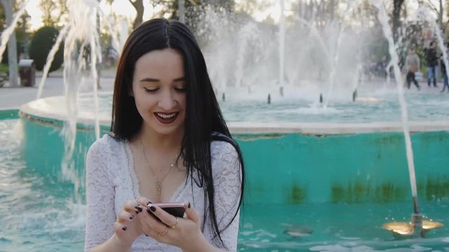Girl laughs using smartphone. Pretty young woman with her mobile phone on fountain background.