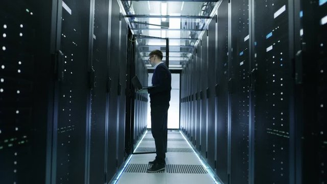 Frontal View of IT Engineer Walking Through Data Center full of Working Rack Servers. He Opens Server Cabinet and Connects to it With His Laptop.  Shot on RED EPIC-W 8K Helium Cinema Camera.