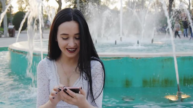 Portrait of girl laughing using smartphone. Pretty young woman with her mobile phone on fountain background.