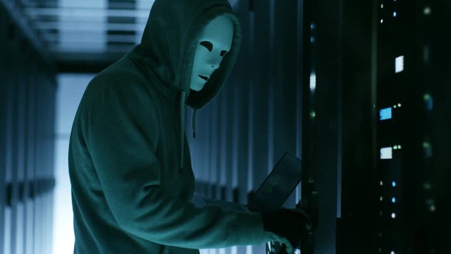 Masked Hacker wearing Hoodie Opens Server Cabinet and Connects to Server with His Notebook.Shot on RED EPIC-W 8K Helium Cinema Camera.