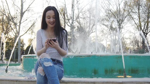 Girl laughs using smartphone. Pretty young woman with her mobile phone on sunset fountain background.