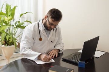 Lekarz w białym kitlu w gabinecie lekarskim siedzi przy stole i wypełnia dokumenty. Lekarz podczas pracy.  - obrazy, fototapety, plakaty