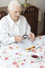 Staruszka siedzi przy stole z gazetą.  Babcia czyta gazetę trzymając szkło powiększające. Babcia czyta artykuł w gazecie za pomocą lupy.