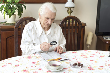 Staruszka siedzi przy stole z gazetą.  Babcia czyta gazetę trzymając szkło powiększające.