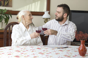 Babcia z wnuczkiem siedzą przy stole i wznoszą toast. Trzymają kieliszki z czerwonym winem. Babcia z wnuczkiem piją wino siedząc przy stole podczas rodzinnego spotkania. - obrazy, fototapety, plakaty