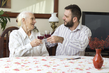 Babcia z wnuczkiem siedzą przy stole i wznoszą toast. Trzymają kieliszki z czerwonym winem.