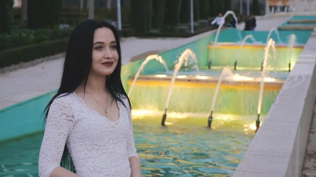 Girl smiles looking and flipping hair. Beautiful young woman on illuminated fountain background.