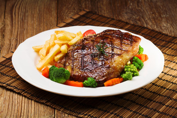 Grilled beef steak served with French fries and vegetables on a white plate.