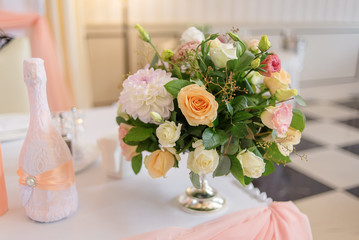 A bouquet of white, cream and pink roses stands in a vase on the table next to a bottle decorated with lace and ribbon