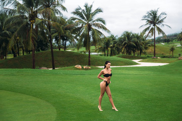 Young sexy and beautiful brunette fitness girl in black swimsuit posing turning booty shows ass at the green grass of Golf club. Sexy model portrait with perfect body. Concept of summer holiday