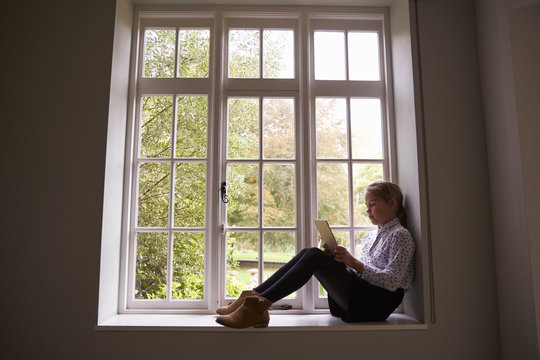 Girl using tablet computer while sitting on window sill at home