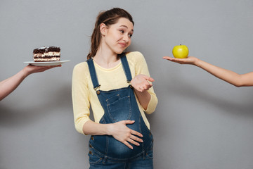 Young confused pregnant woman choosing between apple and cream cake
