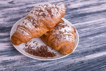 Croissants with powdered sugar and chocolate on white plate on colored wood background. Close up