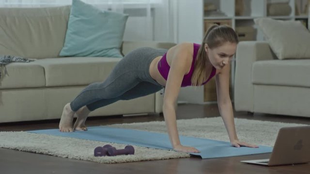 Tracking shot of young fit woman watching workout video tutorial on laptop and doing pushups on mat at home and then stretching her back