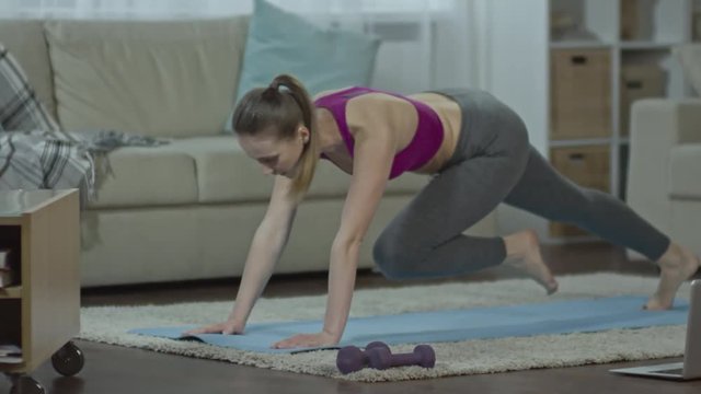 Slow motion shot of woman in sports bra and leggings running in place in a plank position on exercising mat on the floor in the living room