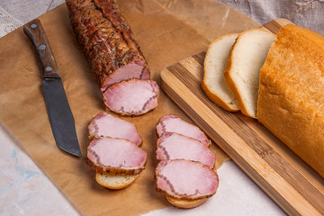 Slices smoked meat or ham on brown packing paper. Slices white wheat bread on wooden cutting board. Small snacks of bread and meat.