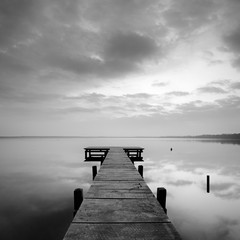 Stiller See mit Steg bei Sonnenaufgang, wolkiger Himmel, schwarz-weiß - obrazy, fototapety, plakaty