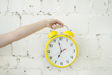 Girl's hand holding yellow alarm clock isolated on white brick backgroung
