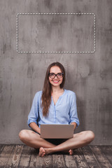 Young smiling woman in glasses sitting on the wooden floor with laptop near grunge grey wall. Copyspace under her head