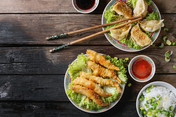 Fried tempura shrimps and gyozas potstickers on lettuce salad with sauces and rice. Served in traditional china plate with chopsticks over old wooden background. Top view. Asian dinner