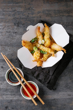 Fried tempura shrimps with rice and spring onion in paper to-go box. Served with sauces, chopsticks, textile napkin over old metal background. Top view, space. Asian take out dinner