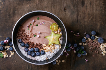 Obraz na płótnie Canvas Smoothie bowl healthy breakfast. Chocolate yogurt with blueberries, granola, coconut, lime and carambola over dark wooden texture background. Drawing branch with flowers. Top view, space