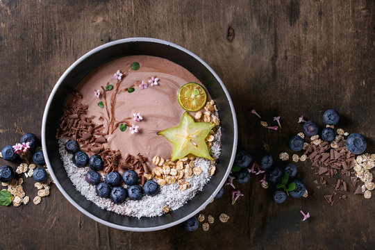 Smoothie bowl healthy breakfast. Chocolate yogurt with blueberries, granola, coconut, lime and carambola over dark wooden texture background. Drawing branch with flowers. Top view, space