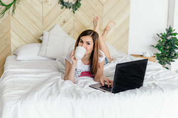 Obraz premium The girl in the bedroom on the bed under white blanket with a laptop and coffee in hand. Business girl. The morning starts with news on the Internet. Lifestyle people concept.