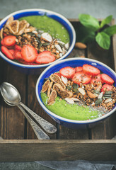 Healthy breakfast. Green smoothie bowls with strawberries, granola, chia and pumpkin seeds, dried fruit and nut in wooden tray over grey concrete background, selective focus. Diet, raw food concept