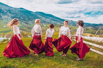 Five beautiful bridesmaids in same color red dress on wedding day having fun and circling around dresses behind mountains and blue sky. Funny girls.