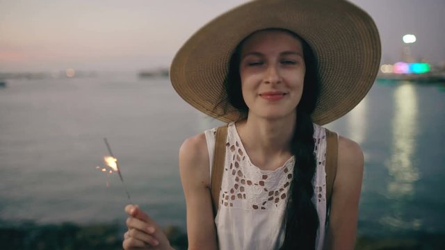 Teenage beautiful girl in hat with burn sparklers and smiling on the beach at sunset