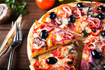 Sliced italian pizza with salami, mushroom, tomatoes, sweet pepper, black olives and purple onion on dark wooden background close up. Italian traditional food. Popular street food.