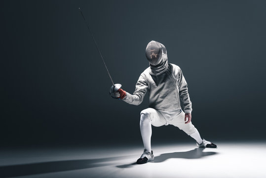 Professional fencer in fencing mask with rapier standing in position on grey