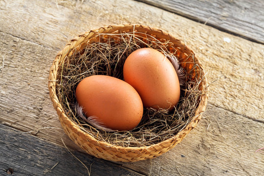 Couple of fresh chicken eggs in a rural basket with hey and feather on a wooden background. Close-up shot. Top view.