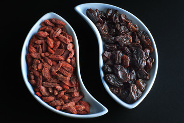 Two curved bowls with dried goji berries and grapes opposite each other on a black background