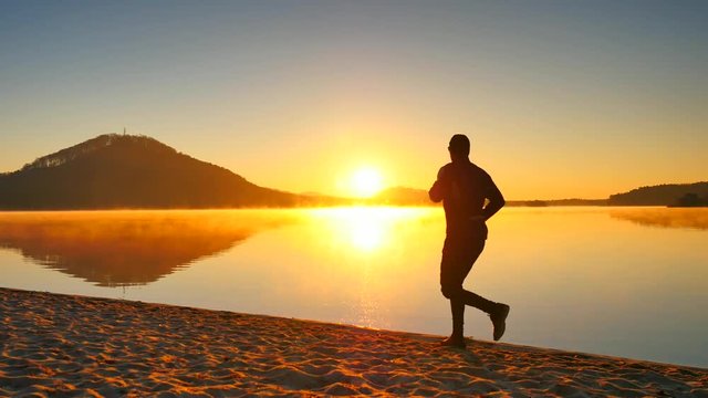 Tall man with sunglasses and dark cap is  running on the beach at sunset.
Silhouette of sport active man running and exercising on the beach of  mountain lake. Orange  sunset sky background.