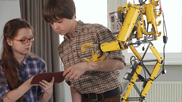Two children checking the toy robot on the table. Pretty girl in glasses holding tablet in her hands. Little brunette boy pointing his hand on their playing device