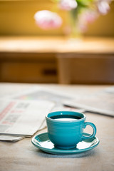 Cup and saucer on table near newspaper with tulips and window light in the background.