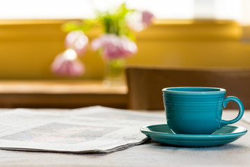 Cup and saucer on table near newspaper with tulips and window light in the background.