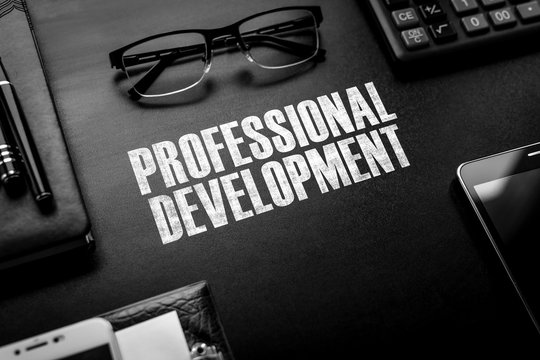 Black chalkboard with business accessories (notepad, magnifying glass, fountain pen, tablet, phone, glasses and calculator) and text PROFESSIONAL DEVELOPMENT. Top view.