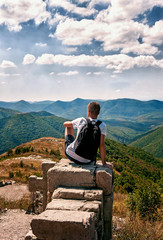 silhouette of man with backpack sitting with his back to the photographer on the big rock and looking at the panorama of mountains , sky and clouds