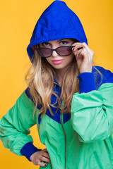 portrait of beautiful blond woman in sunglasses and blue green hooded jacket on yellow background. hipster summer.