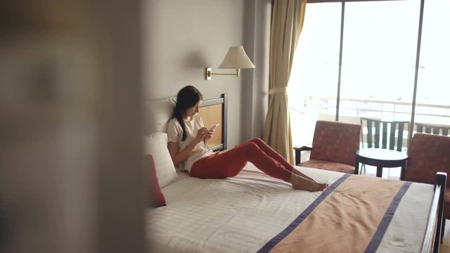 Pretty blonde woman lying in bed in hotel room and use smartphone and smiling
