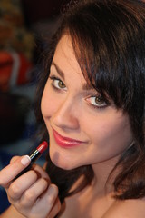 Obraz premium Lovely young brunette applying lipstick, against a darkened room.