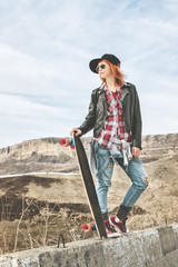 Naklejka premium Stylish red-haired girl in a cap and denim overalls standing on a bump at the highway in the mountains with a longboard