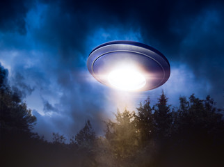 High contrast image of UFO flying over a forest with light beam at night