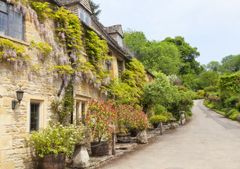 Fototapeta na wymiar Old english cottages with flowering purple wisteria on the walls, stone mushroom ornaments, by a road, in a rural countryside village