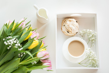 Morning breakfast in spring with a cup of black coffee with milk and pastries in the pastel colors, a bouquet of fresh yellow and pink tulips on a white background. Top view.