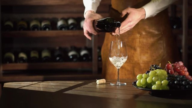 The nice composition of the bundles of the juicy colourful grapes on the black plate and the cork on the table. The sommelier is filling the glass with the white tasty wine.