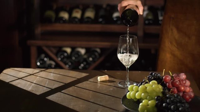 The close-up portrait of the somiellier pouring the white wine into the glass at the background of the shelves full of wine. The composition of the colourful graped on the black plate.